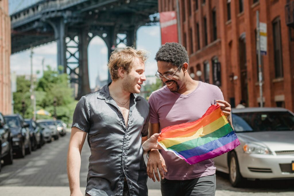 Happy interracial same-sex couple celebrates LGBT pride with a rainbow flag on an urban street.