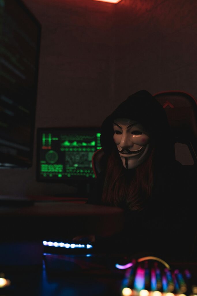 An anonymous person wearing a Guy Fawkes mask working on a computer in a dark room, suggesting cyber activities.