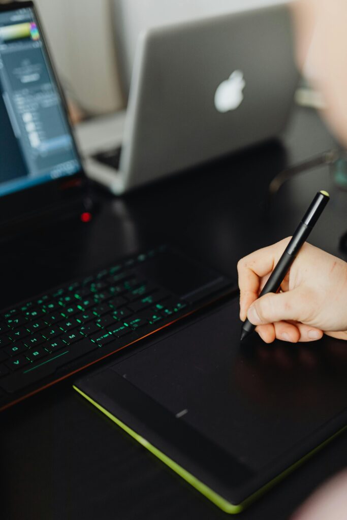 Hand holding stylus pen writing on digital tablet with laptop setup, perfect for creative workspaces.